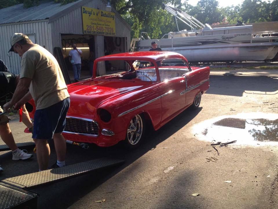 Red classic car on a ramp, people nearby. Building and boat in background.