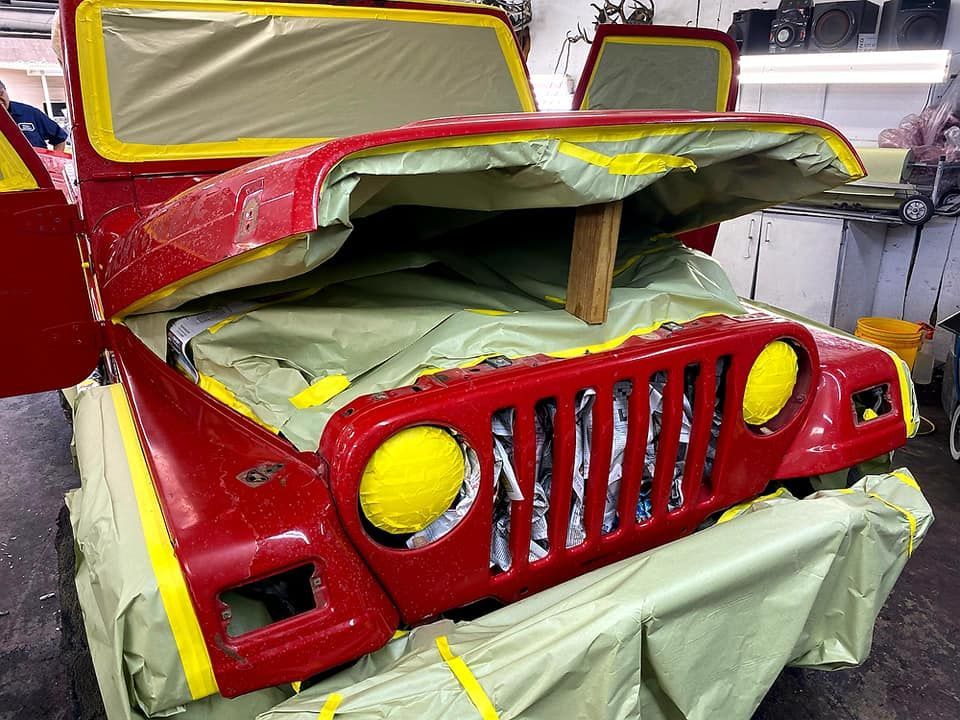 Red Jeep being prepped for painting, masked off with yellow tape and green plastic in a garage.
