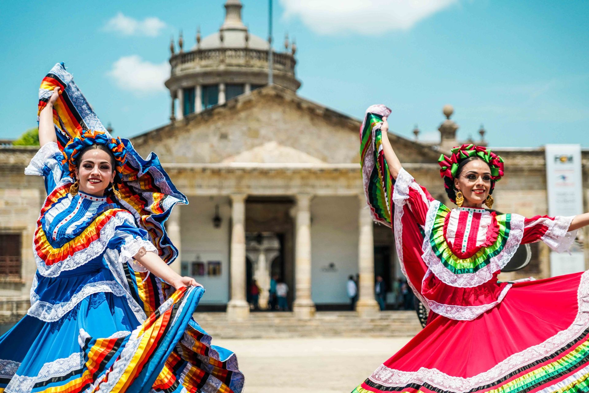 Dos bailarinas con coloridos trajes folclóricos actuando frente a un edificio histórico de piedra.