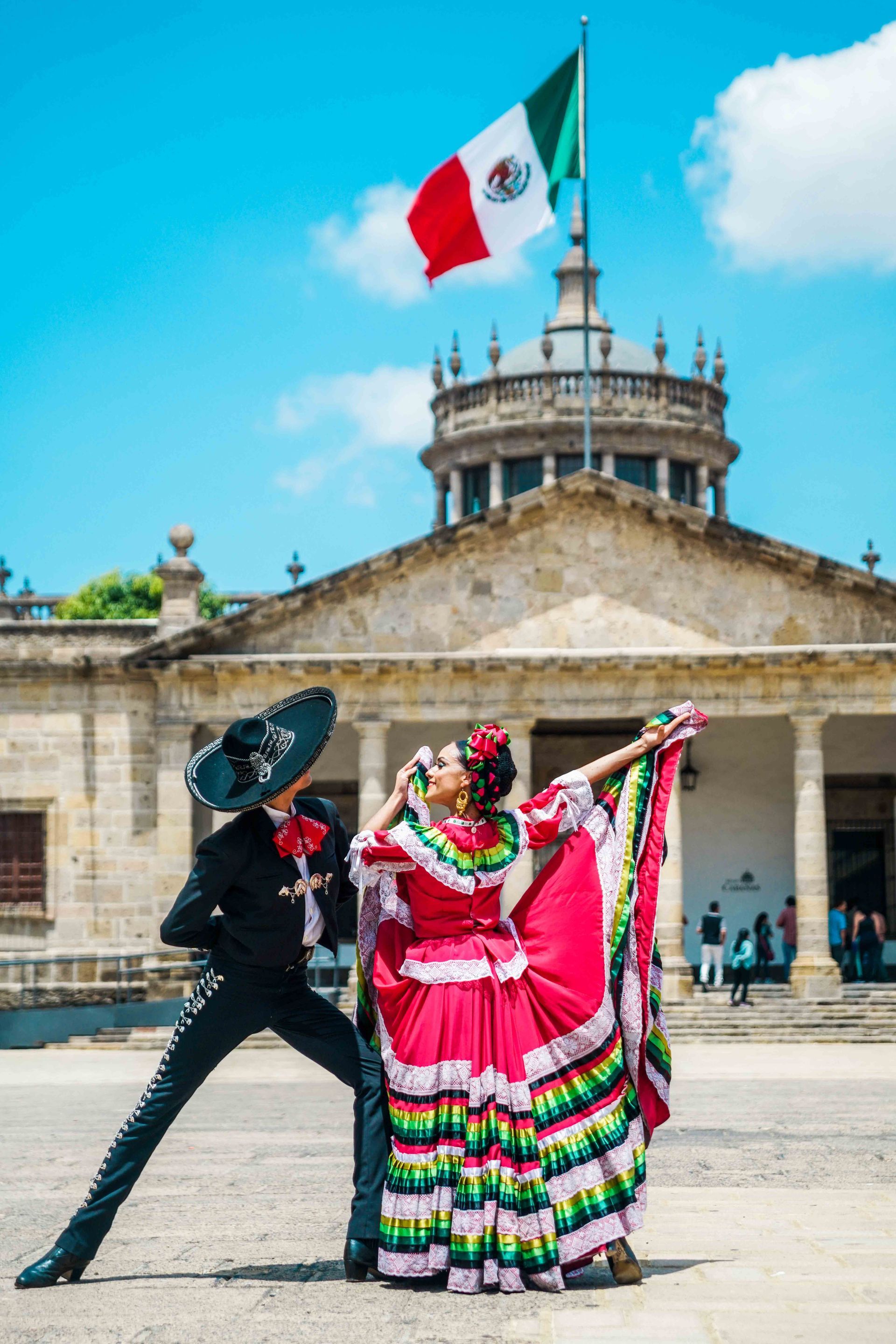 Bailarines con coloridos trajes folclóricos actúan frente a un edificio histórico con una bandera mexicana.