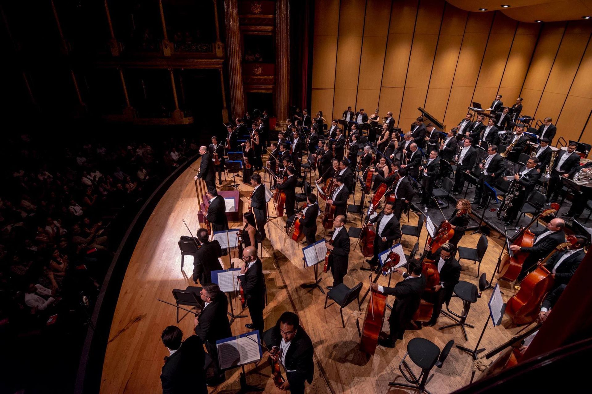 Orquesta actuando en el escenario de una sala de conciertos bajo una iluminación cálida, con los músicos sentados en filas.