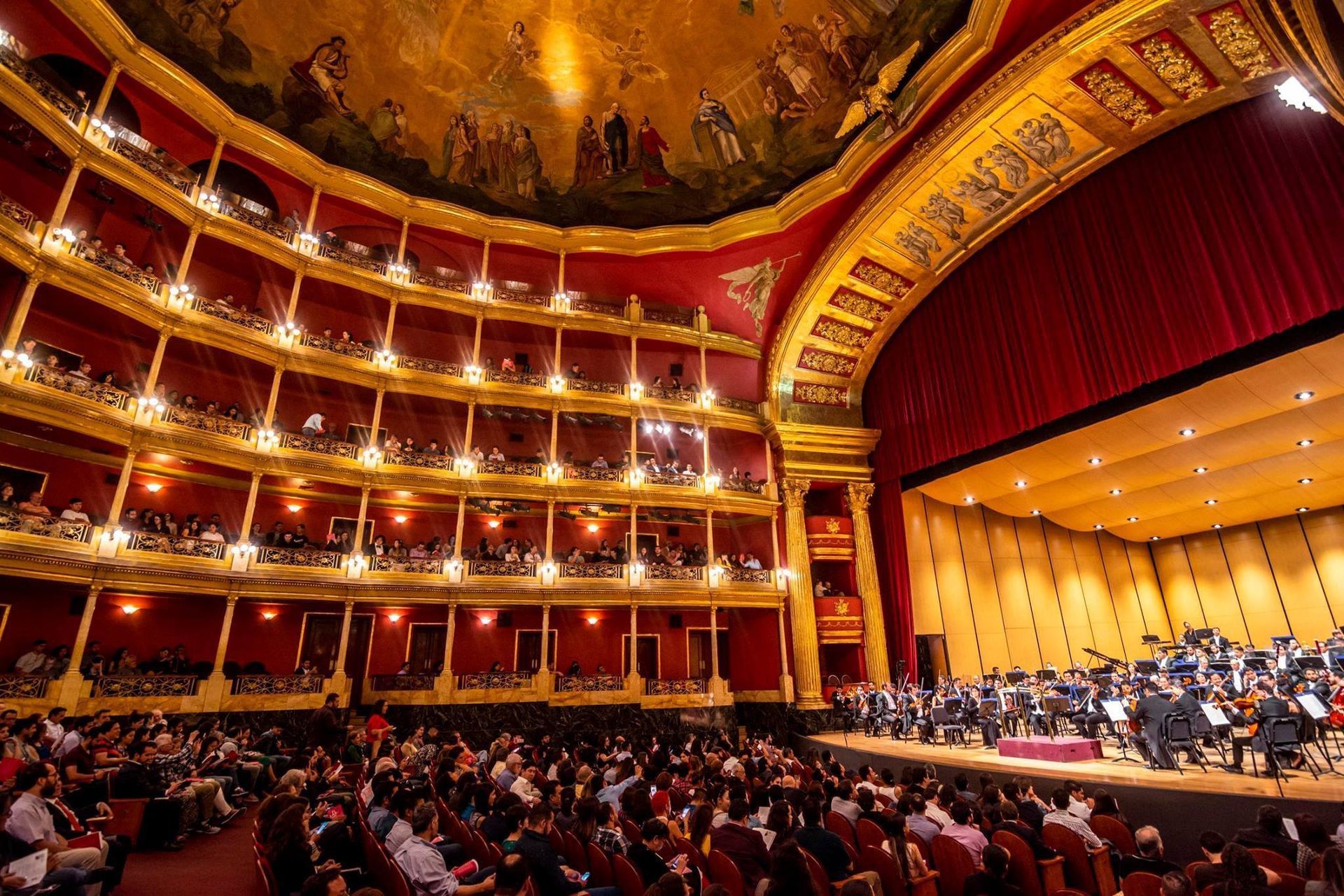 Elegante auditorio con butacas rojas y una orquesta iluminada en el escenario bajo un techo pintado.