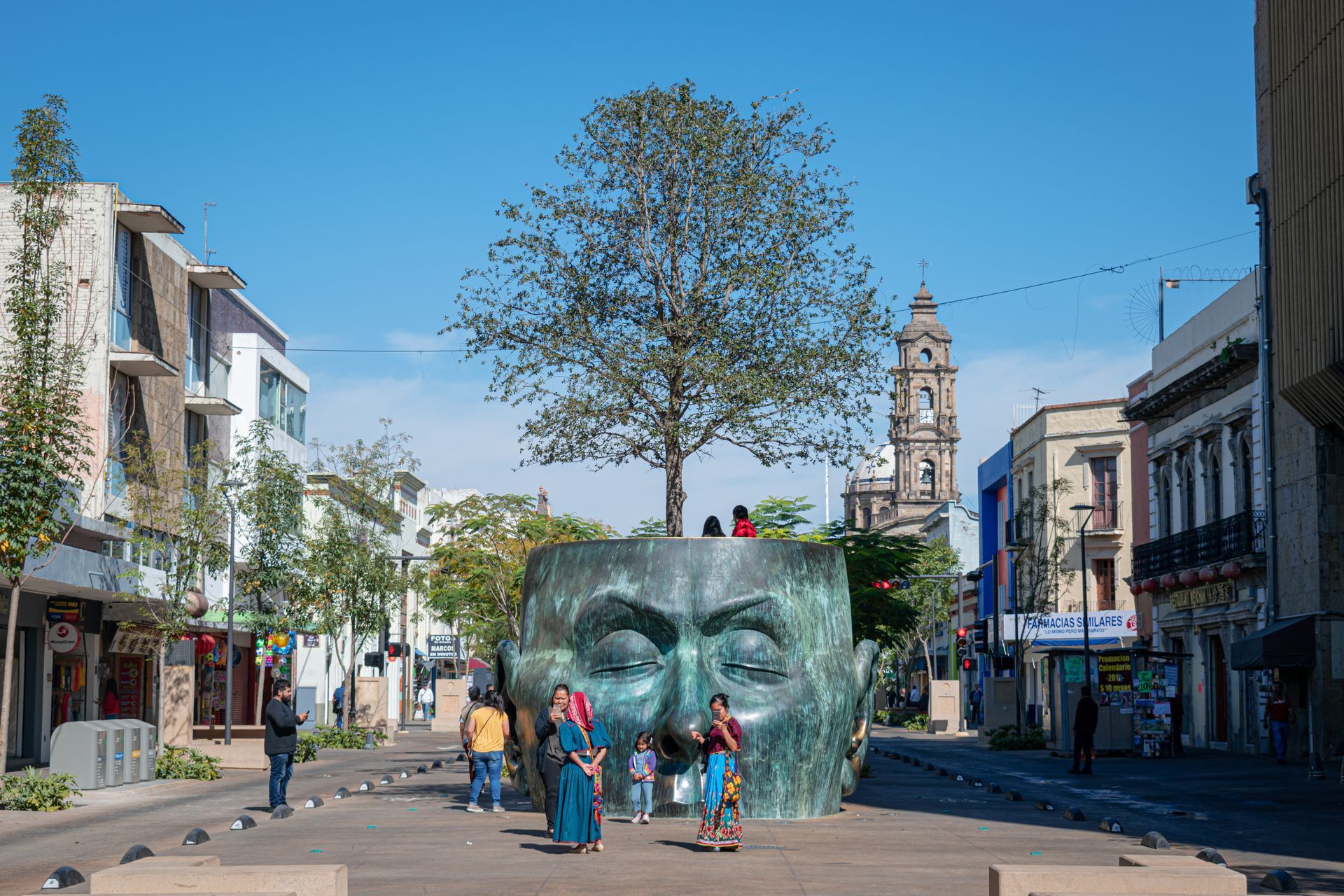 Personas reunidas alrededor de una gran escultura de un rostro en una plaza soleada, con edificios y una torre al fondo.