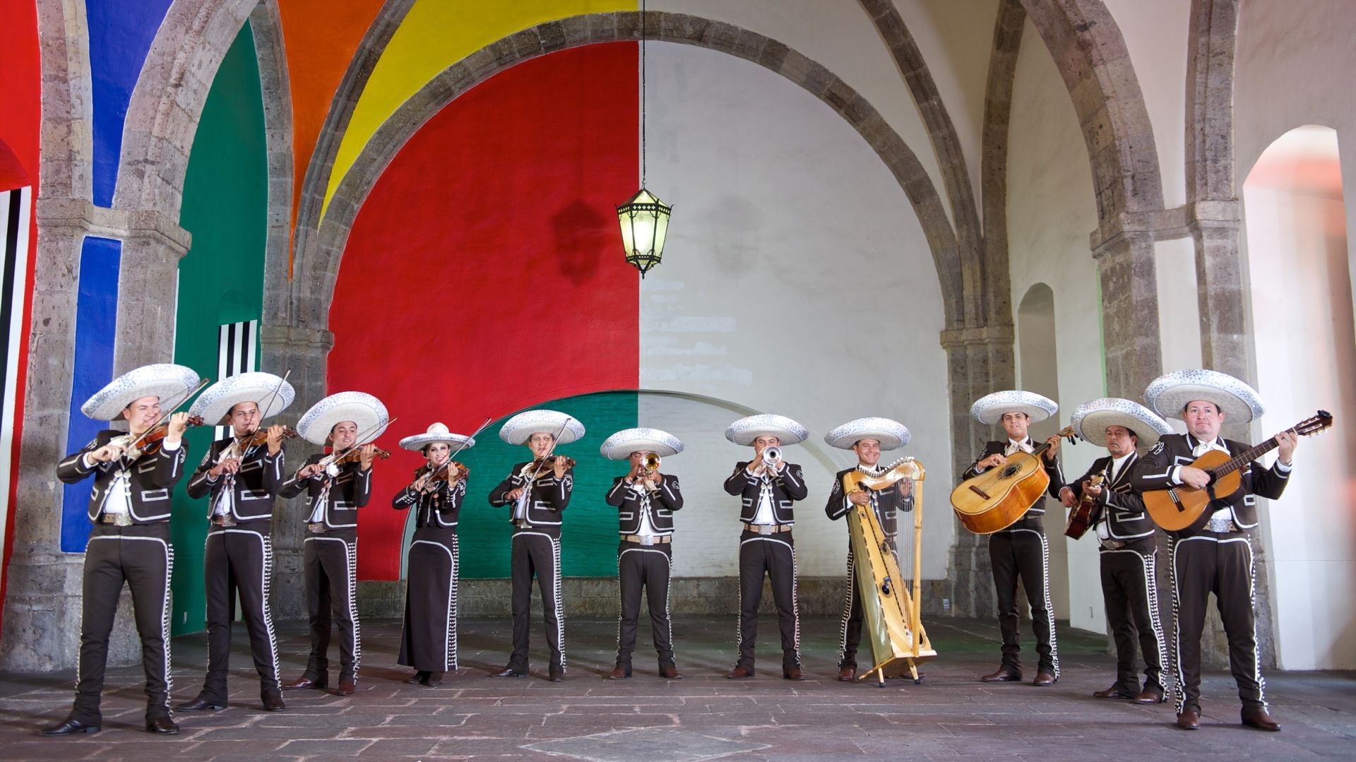 Grupo de mariachis con sombreros actuando frente a coloridos arcos y una pared roja y blanca.
