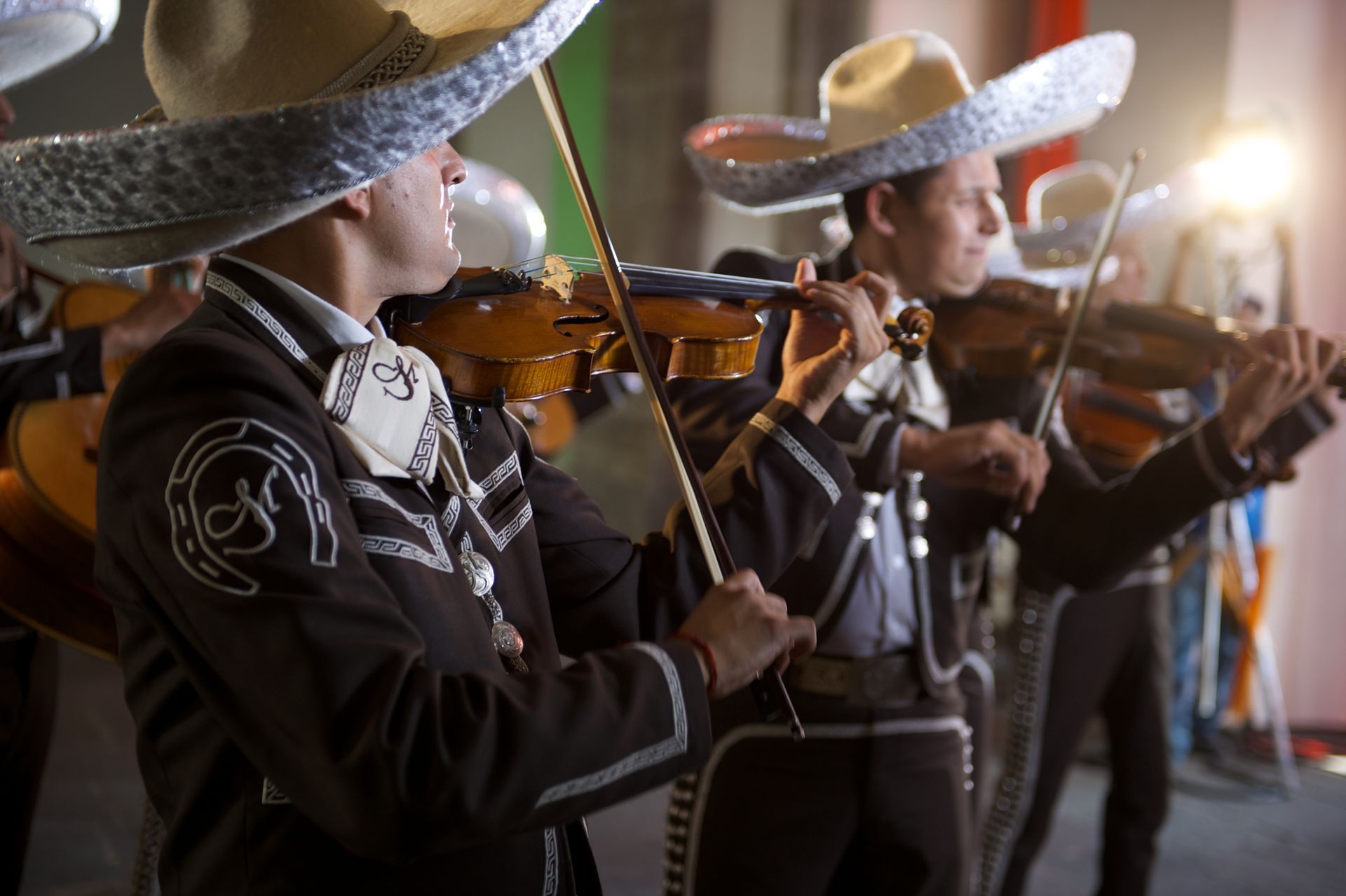 Músicos de mariachi con sombreros tocando violines durante una actuación en vivo en un espacio cerrado.