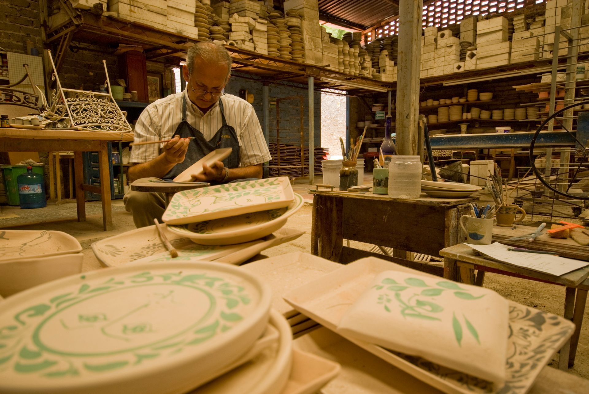 Persona pintando platos de cerámica en un taller, con pilas de platos decorados en primer plano.