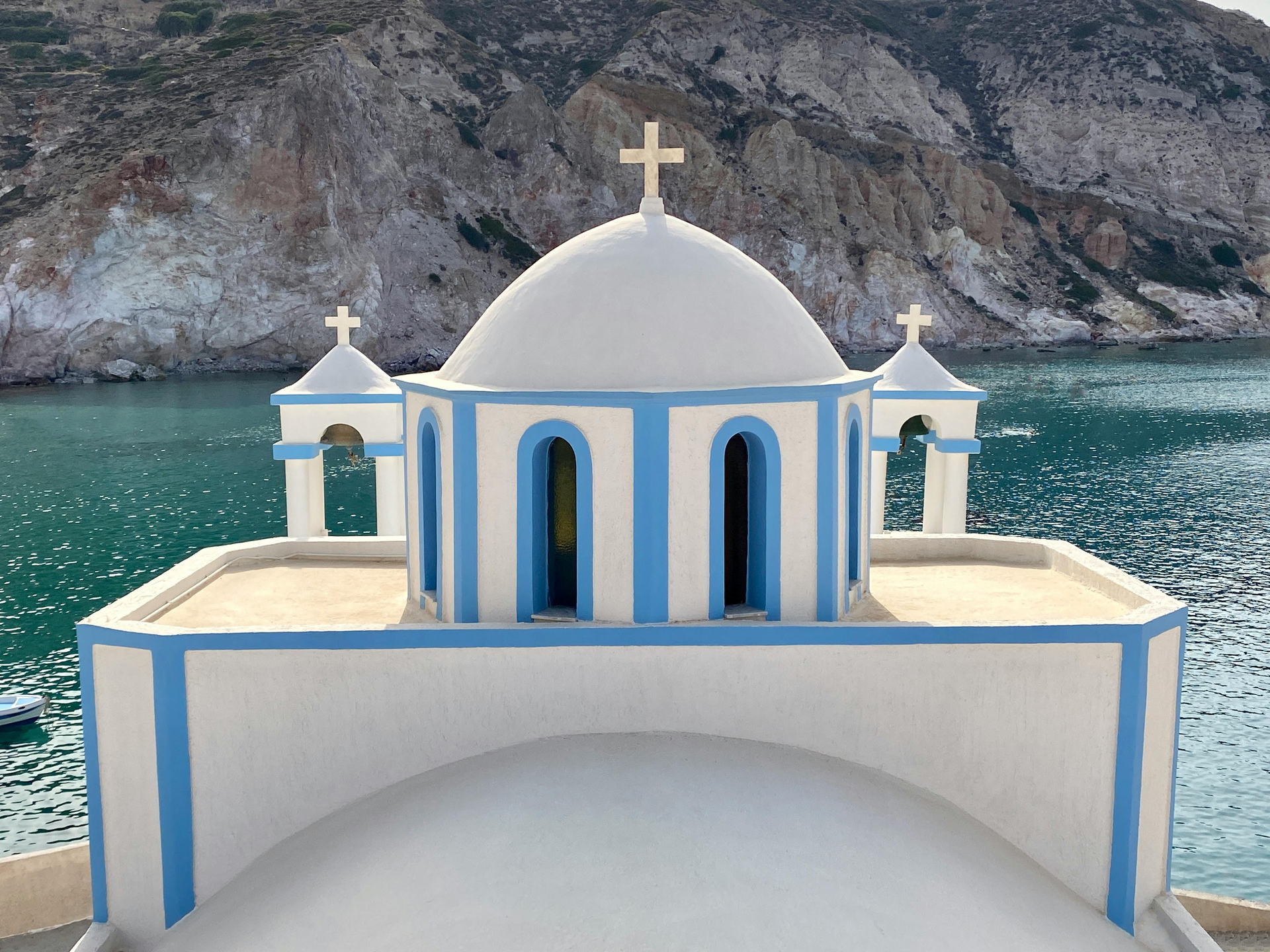 Capilla costera blanca y azul con techo abovedado y cruz, con vistas a aguas turquesas y acantilados rocosos.