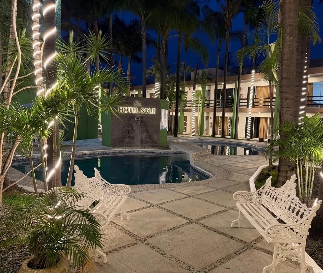 Patio de piscina iluminado por la noche, con palmeras, bancos blancos y un edificio iluminado al fondo.