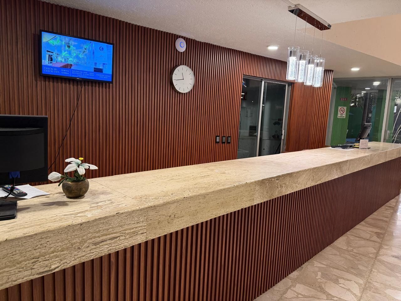Mostrador de recepción con pared de madera, mostrador de granito, reloj, televisor y ascensor.
