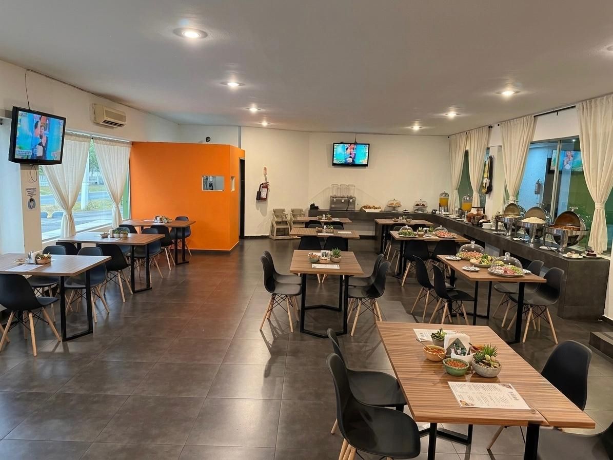 Comedor luminoso con mesas preparadas para invitados, sillas negras y una pared decorativa naranja bajo luces empotradas.