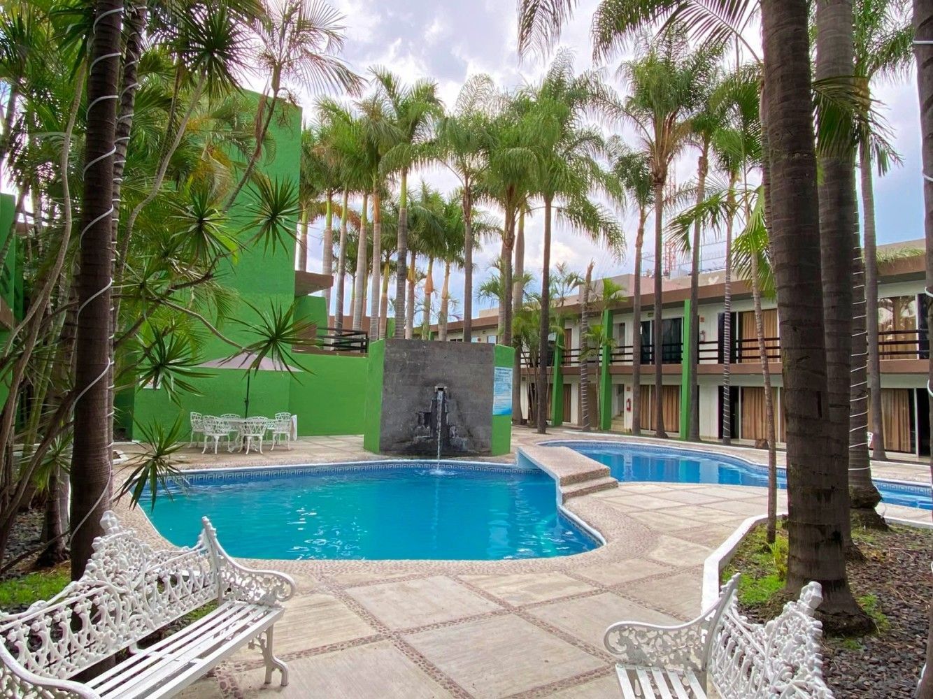 Piscina de un complejo turístico tropical con palmeras, tumbonas blancas y un edificio verde al fondo.