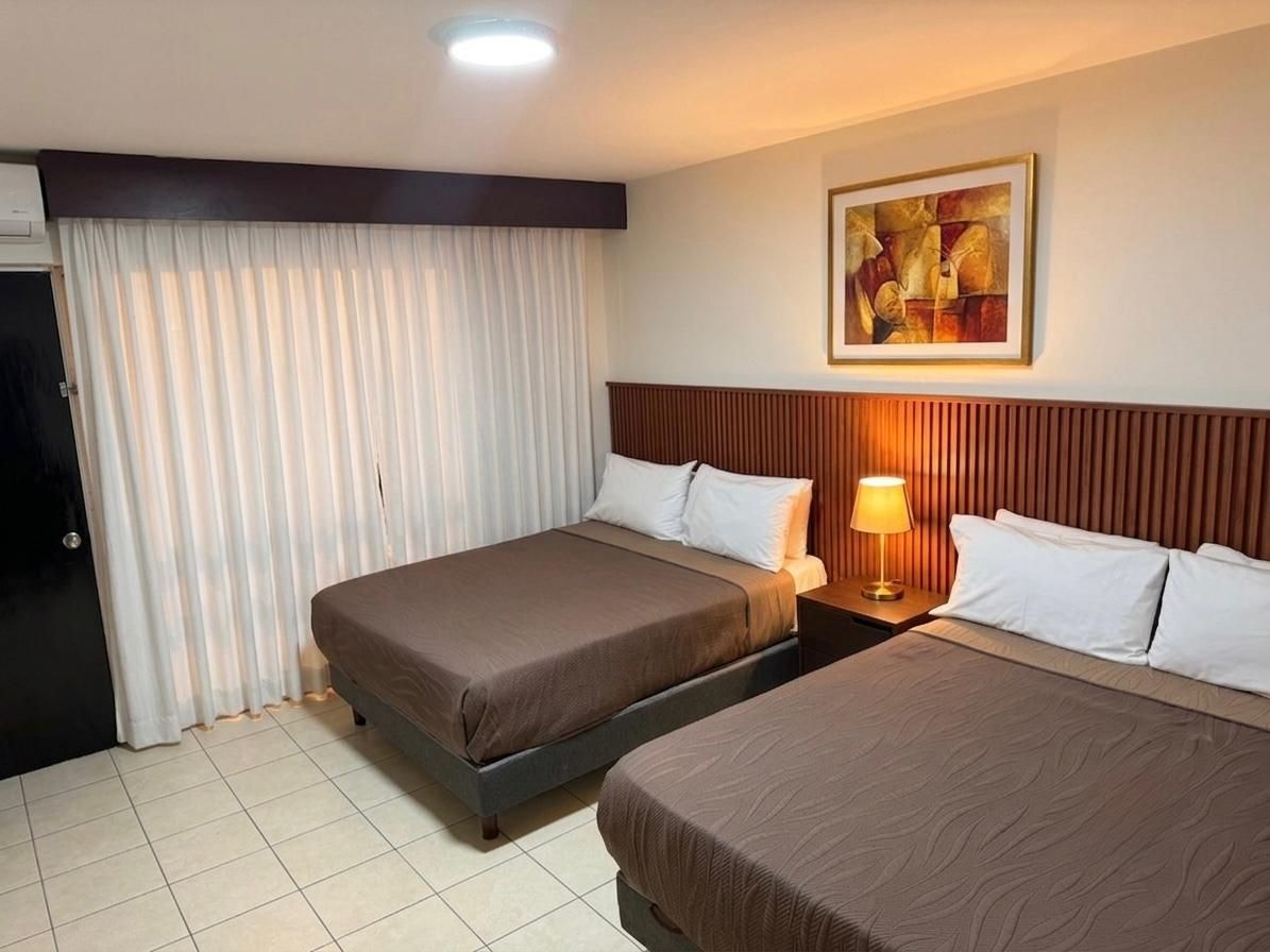 Habitación de hotel con dos camas, lámpara de noche, cuadro en la pared y grandes cortinas correderas.