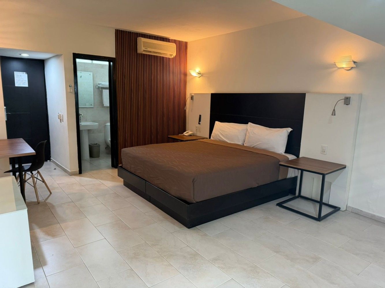 Habitación de hotel moderna con una cama grande, aire acondicionado de pared, puerta de baño y un pequeño escritorio.