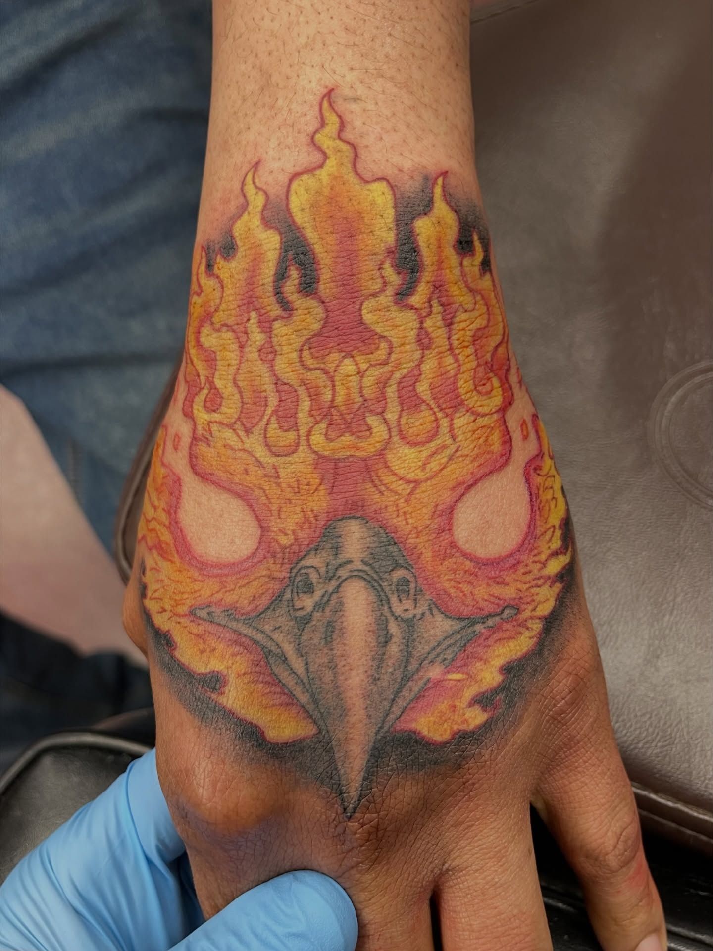 Colorful flame-themed tattoo on the back of a hand and wrist, with bold orange, yellow, black, and pink details