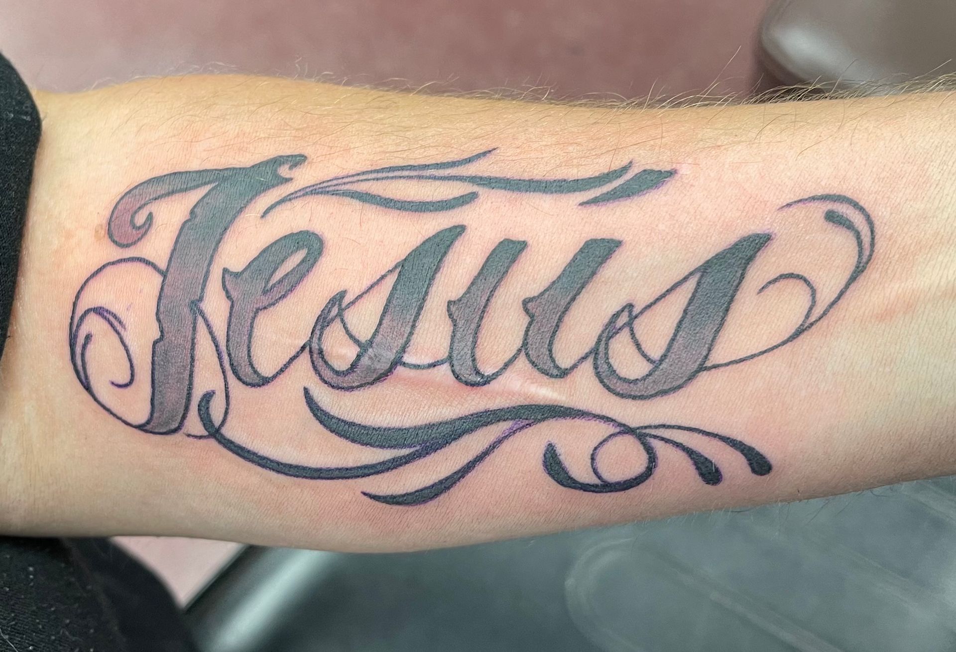 Forearm tattoo reading “Jesus” in black cursive with blue shading and decorative flourishes