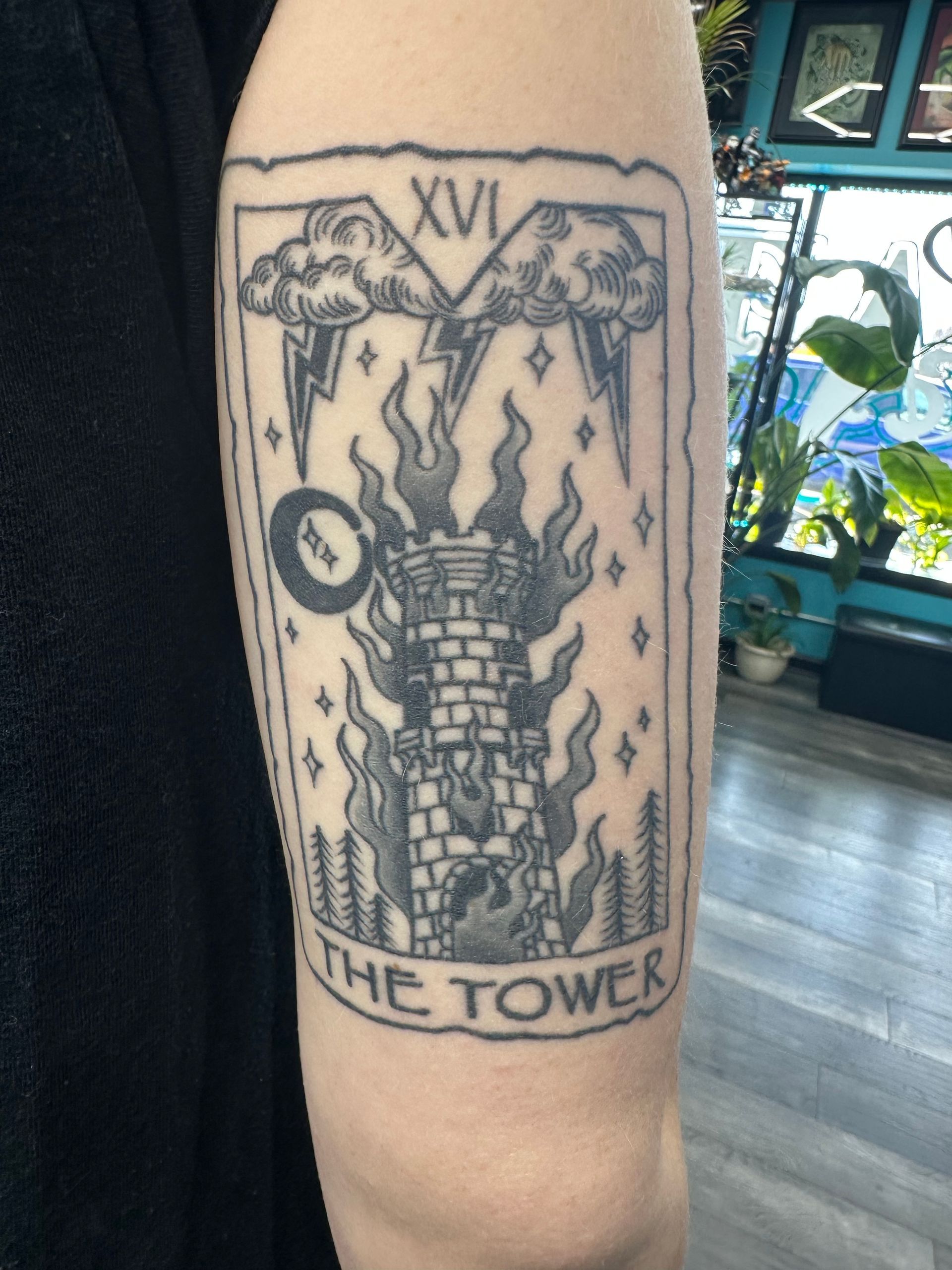 Forearm tattoo of a tarot card labeled “XV” and “The Tower,” showing a burning tower with lightning.