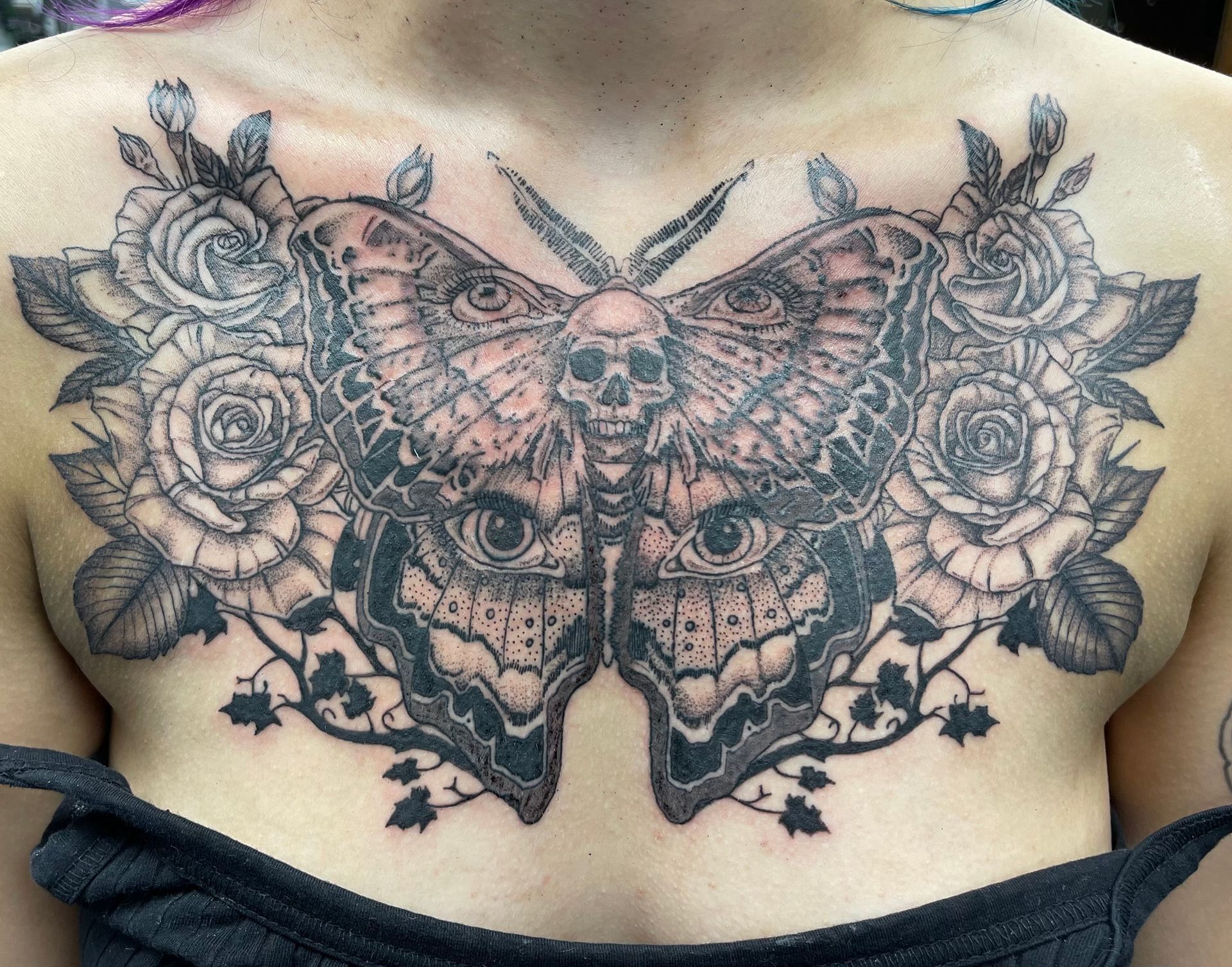 Large black-and-gray butterfly and skull tattoo across the upper back, framed by roses and floral details