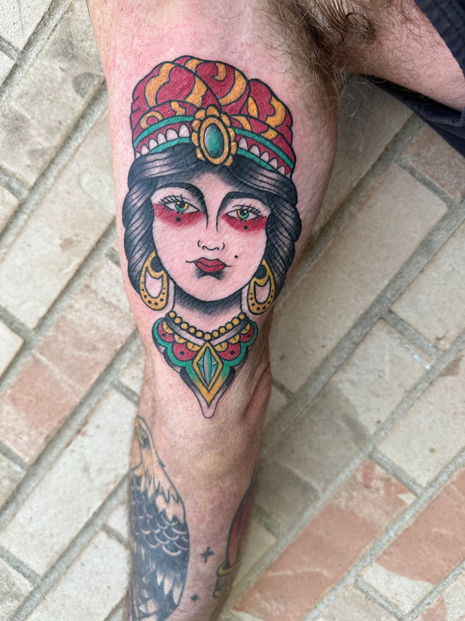 Colorful traditional-style face tattoo on a person’s calf, with ornate teal, red, and gold details.