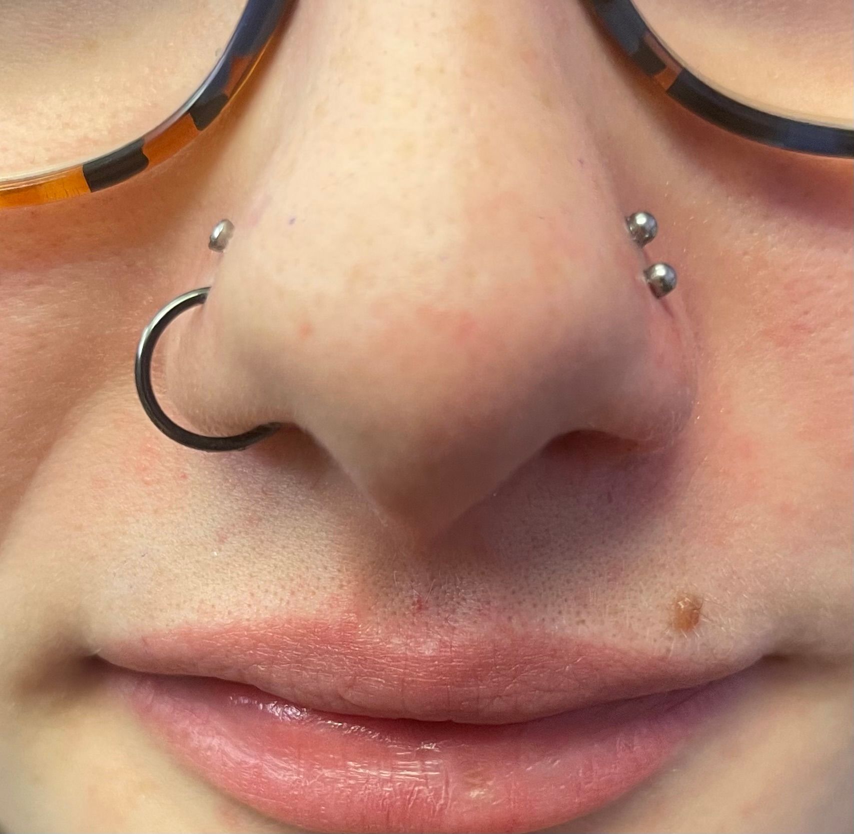 Close-up of a nose with a hoop and stud piercings, framed by glasses and a small mole above the lip