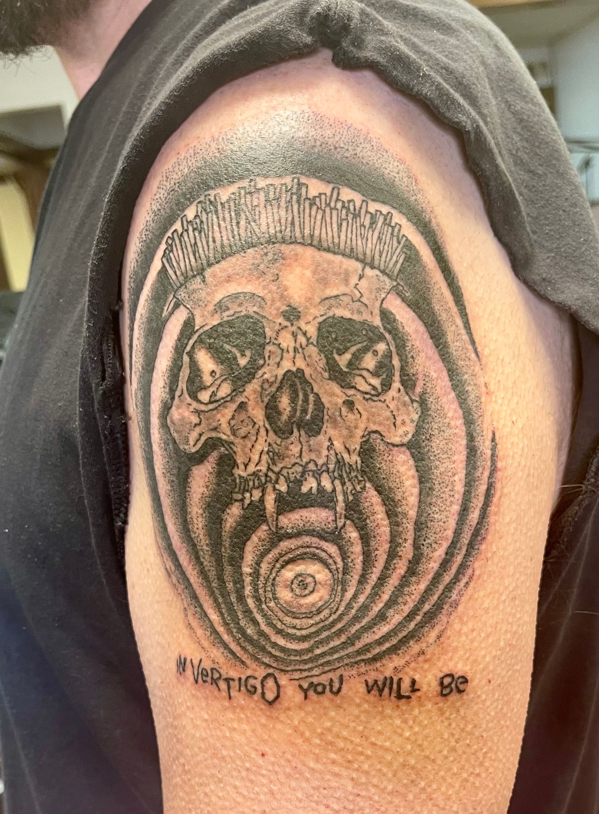 Tattoo of a skull-faced figure in a headdress on a person’s upper arm, with text below.