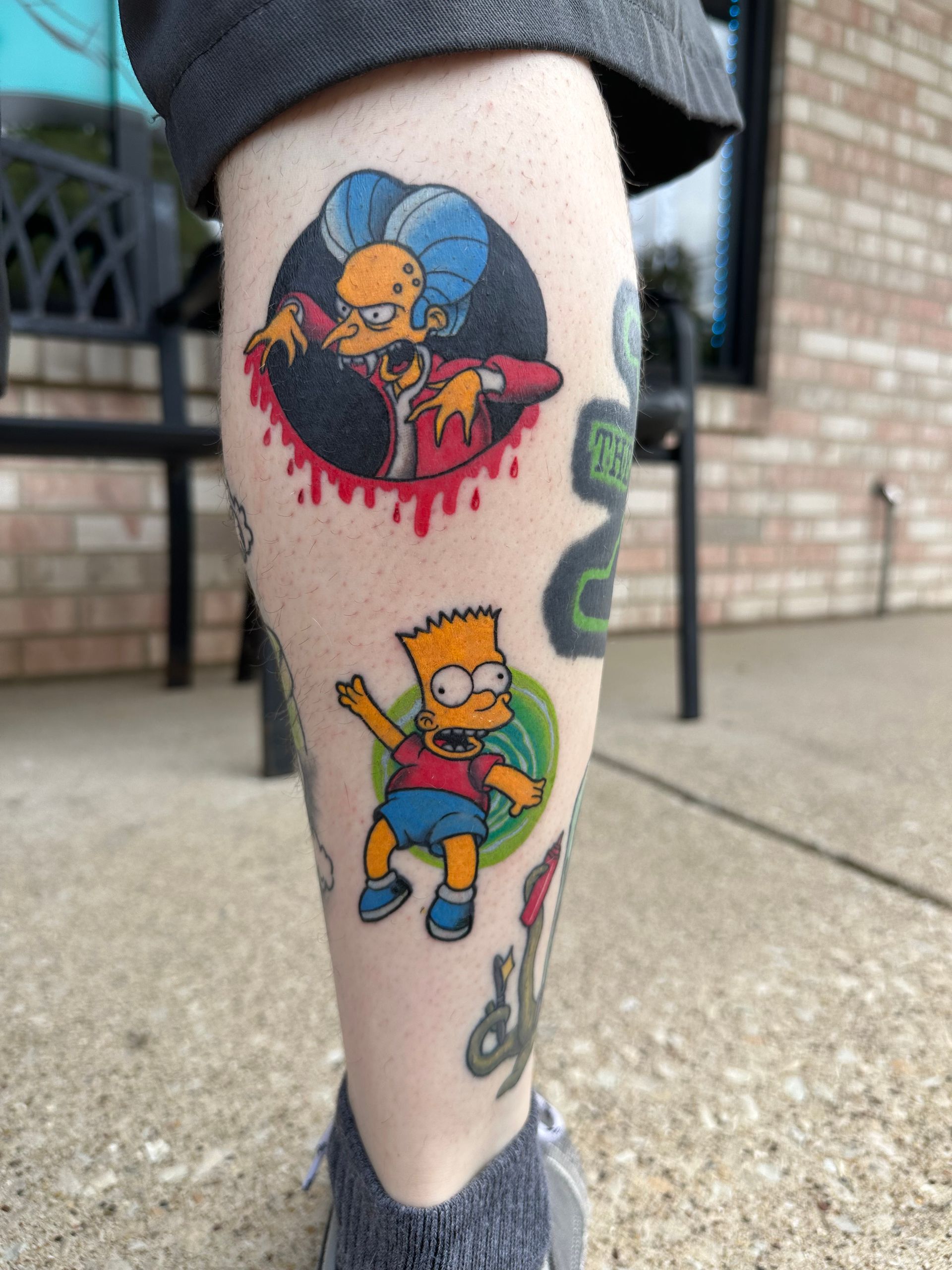 Two colorful cartoon tattoos on a lower leg near a brick wall and sidewalk
