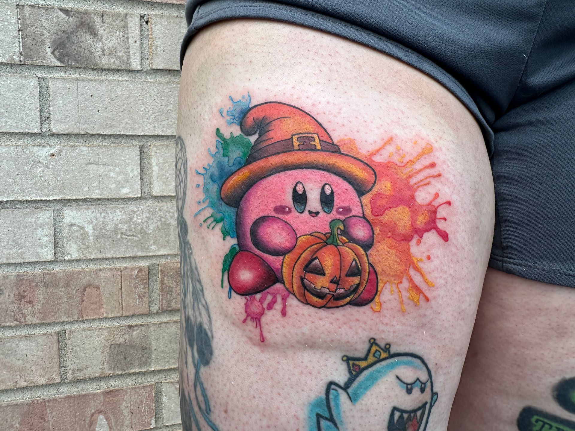 Kirby Halloween tattoo with a jack-o’-lantern, pink splash background, and small cartoon shark below