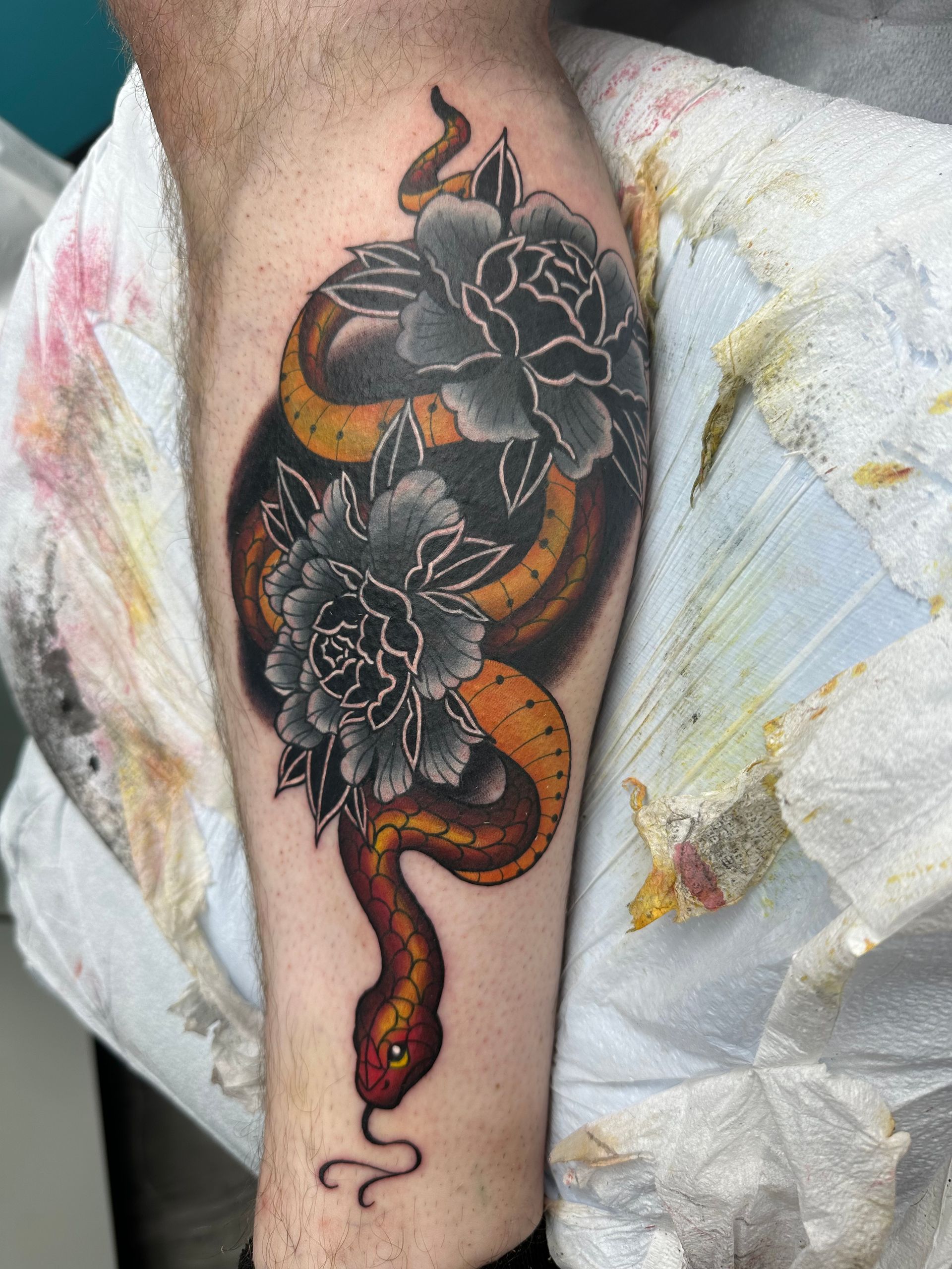 Black and orange snake tattoo with large flowers on a calf, partially covered by a cloth.