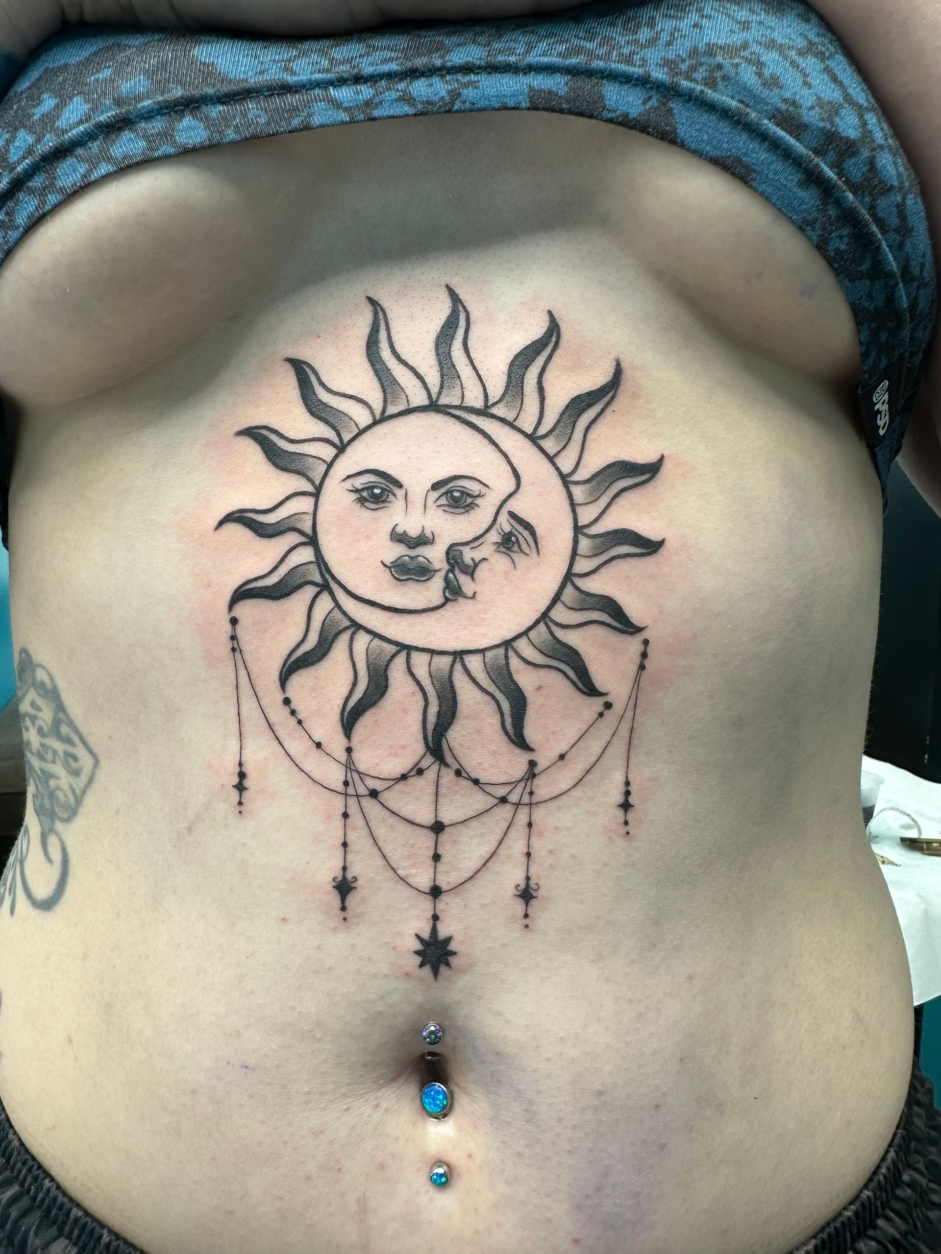 Torso with sun-and-moon tattoo and dangling belly jewelry, wearing blue patterned clothing above the waist