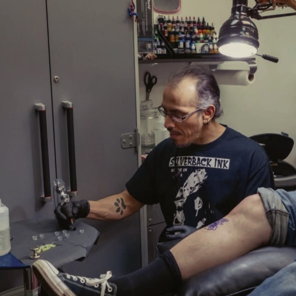 Tattooed man getting a tattoo in a studio, seated under a bright lamp while an artist works on his arm.