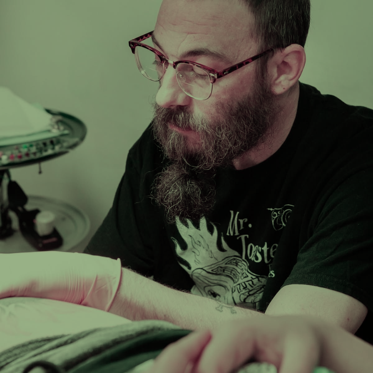 Man with glasses and beard reading an open book at a table in soft green light