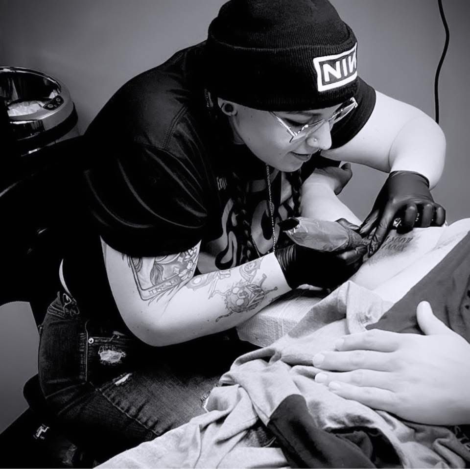 Tattoo artist in black cap and gloves working on a client’s arm in a black-and-white studio scene