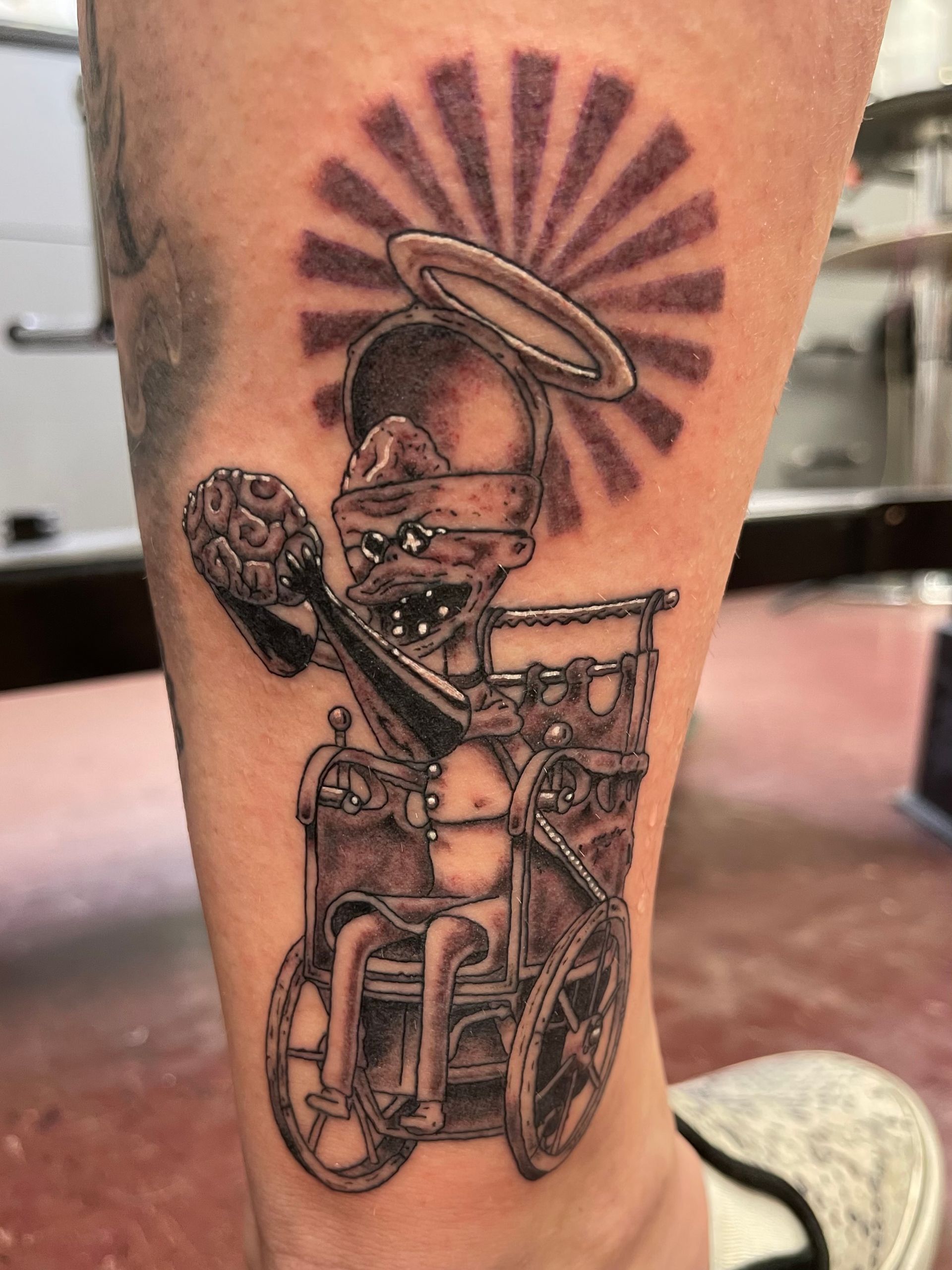 Black-and-white tattoo of a skeleton in a top hat with a halo, seated in a wheelchair with a raised arm