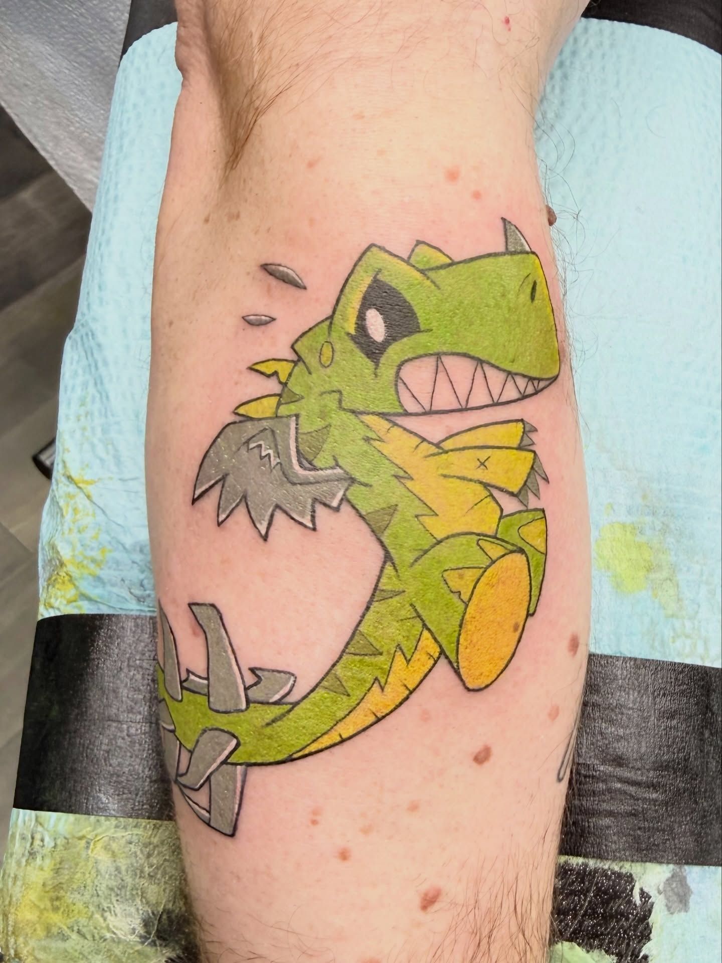 Colorful cartoon dragon tattoo on a calf, featuring green, yellow, pink, and gray with a wide grin.