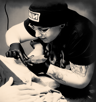Tattoo artist in black gloves working on a client’s arm, leaning close in a monochrome scene
