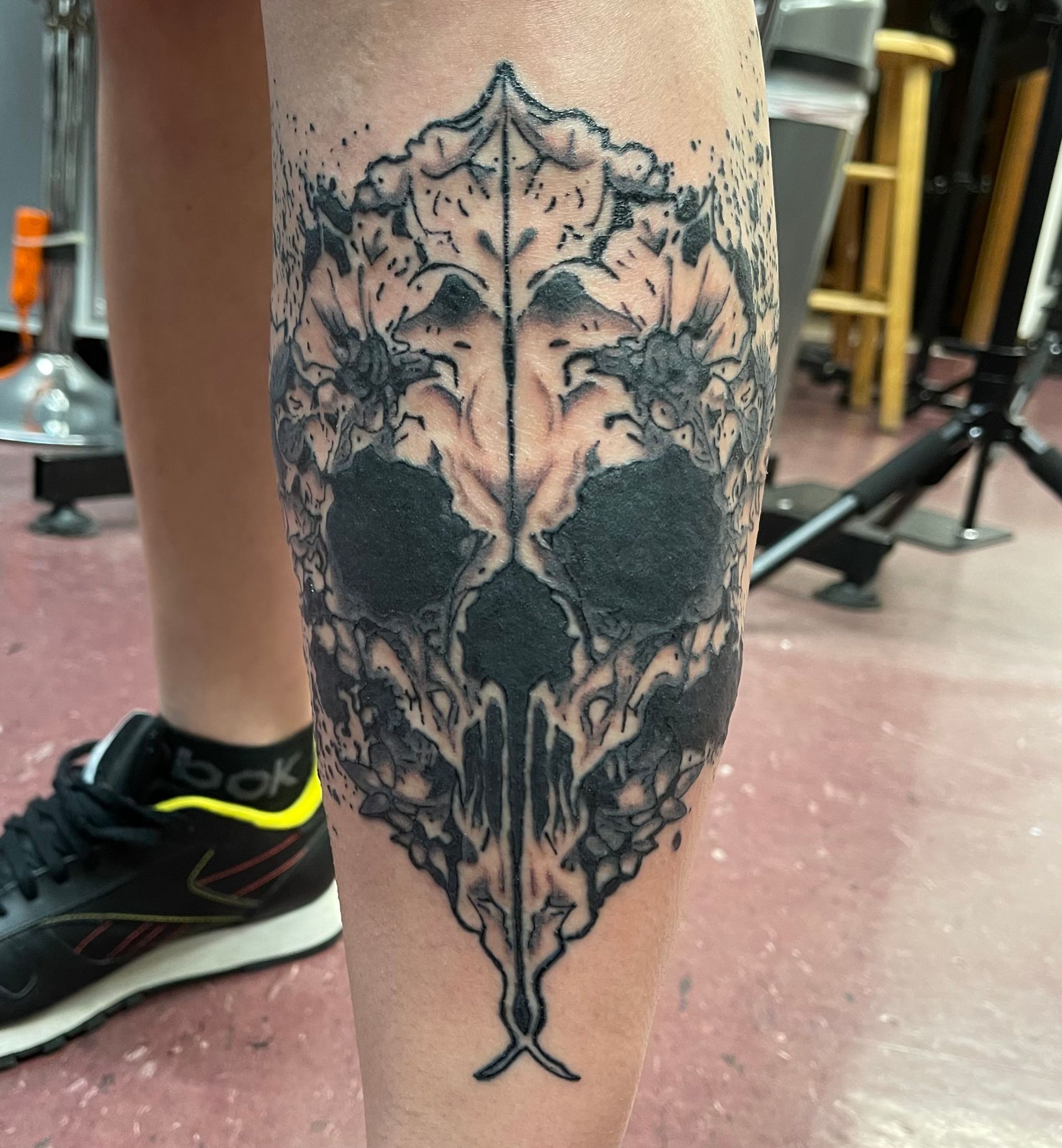 Close-up of a black skull tattoo on a calf in a gym setting with a sneaker and equipment in the background
