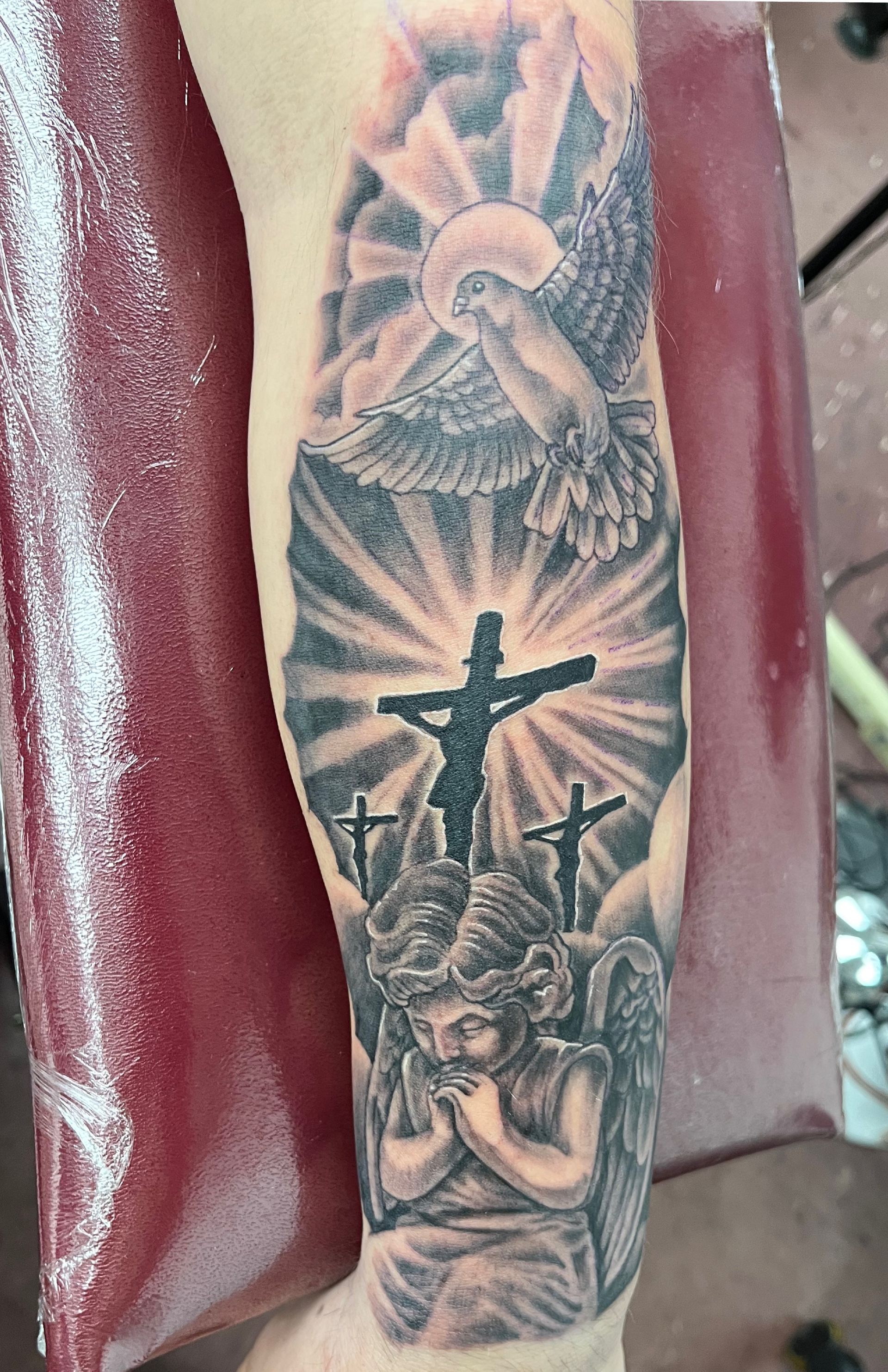 Black-and-gray religious tattoo with cross, praying figure, rays, and angel on a forearm