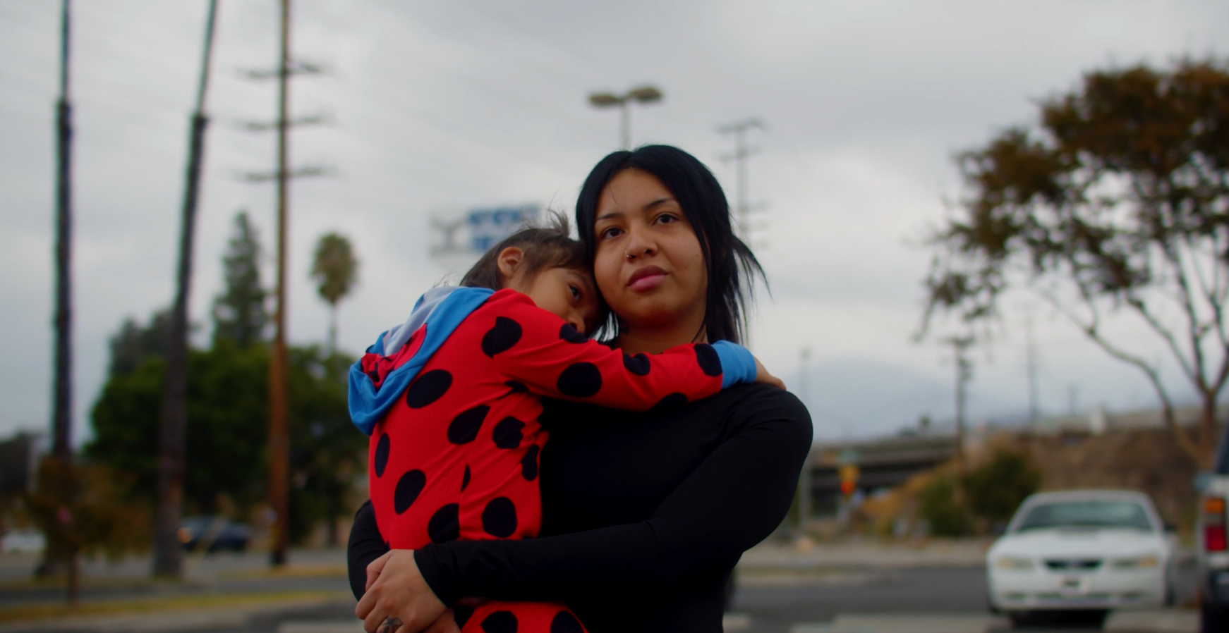 Woman holding a child outdoors; cloudy sky, parking lot background.
