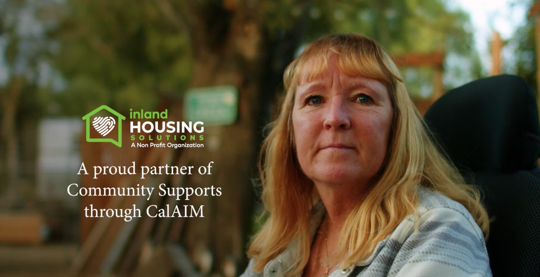 Woman in wheelchair, blonde hair, looking forward. Outdoor setting. Inland Housing logo, 