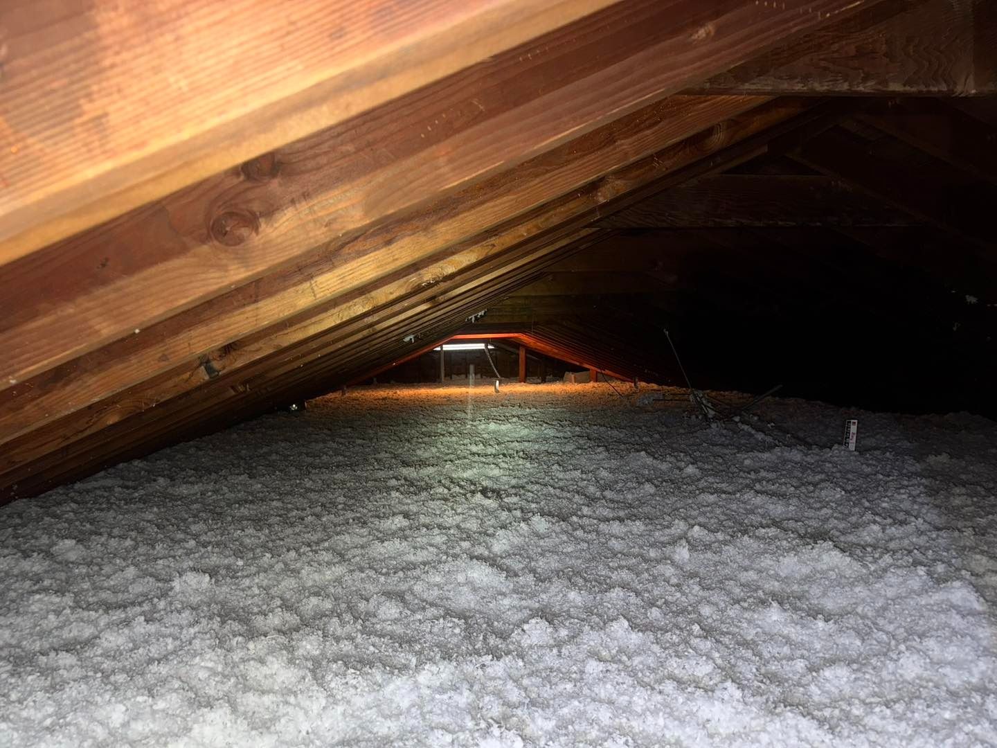Attic filled with white insulation, illuminated by a light, under wooden rafters.