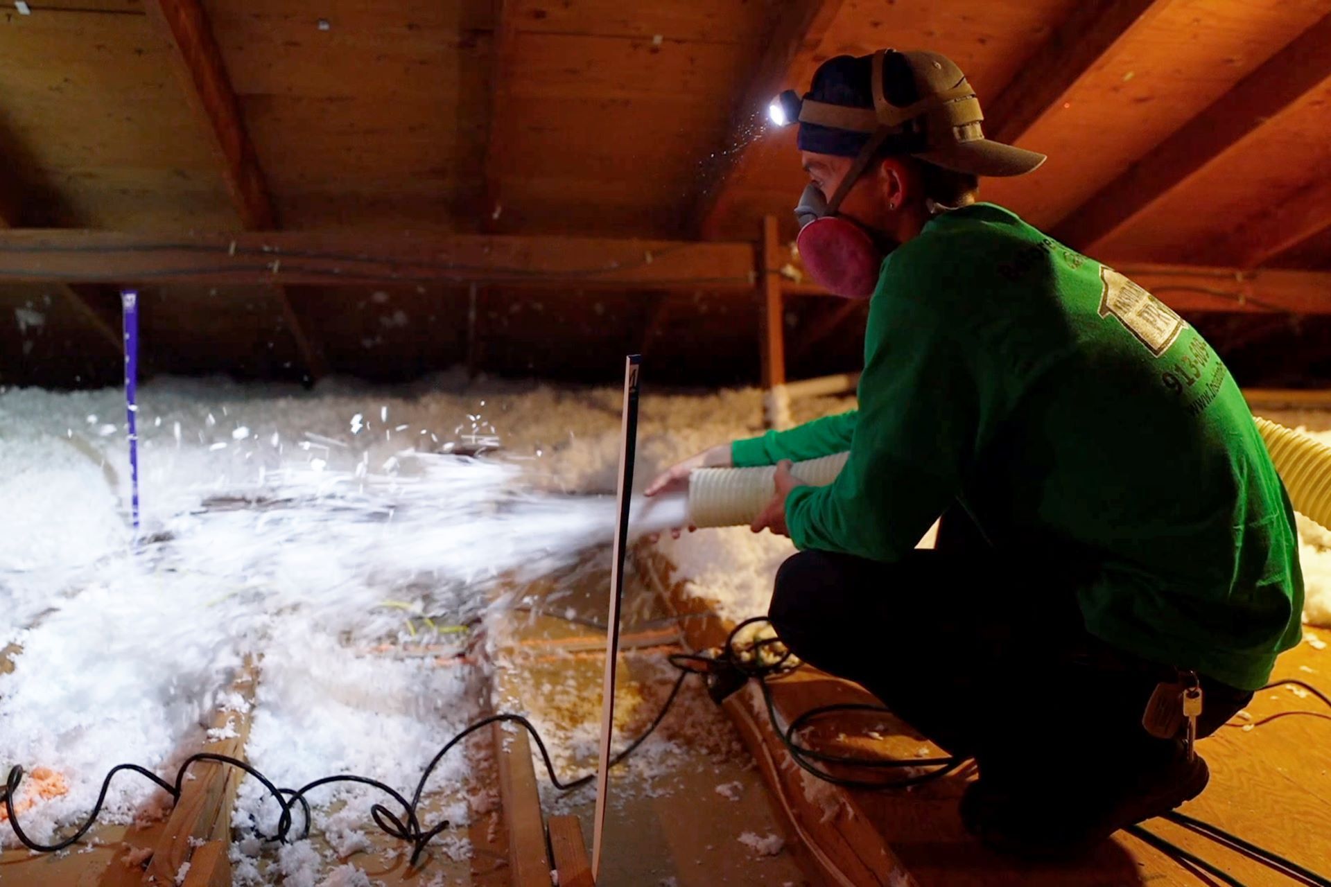Worker in green jacket and helmet inspecting insulation in an attic crawlspace with spray foam and tools.