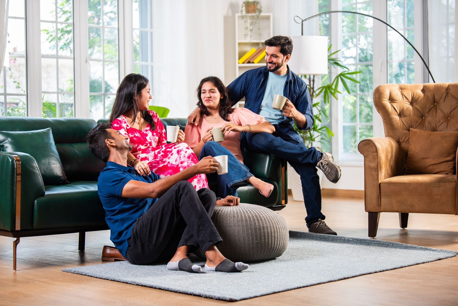 Four people relaxing in a living room, some sitting on a green couch, others on the floor, holding mugs, smiling.