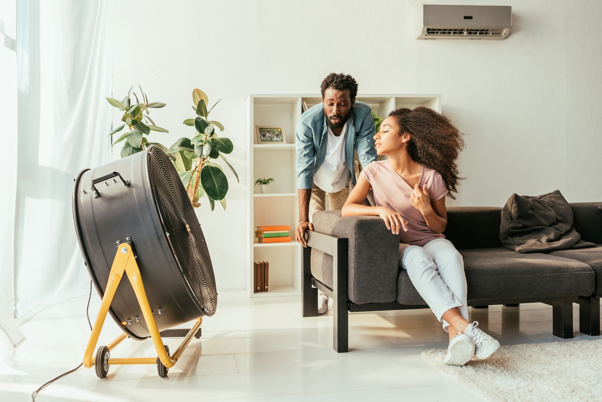 Man and woman in a bright living room moving a large black fan on a tripod stand