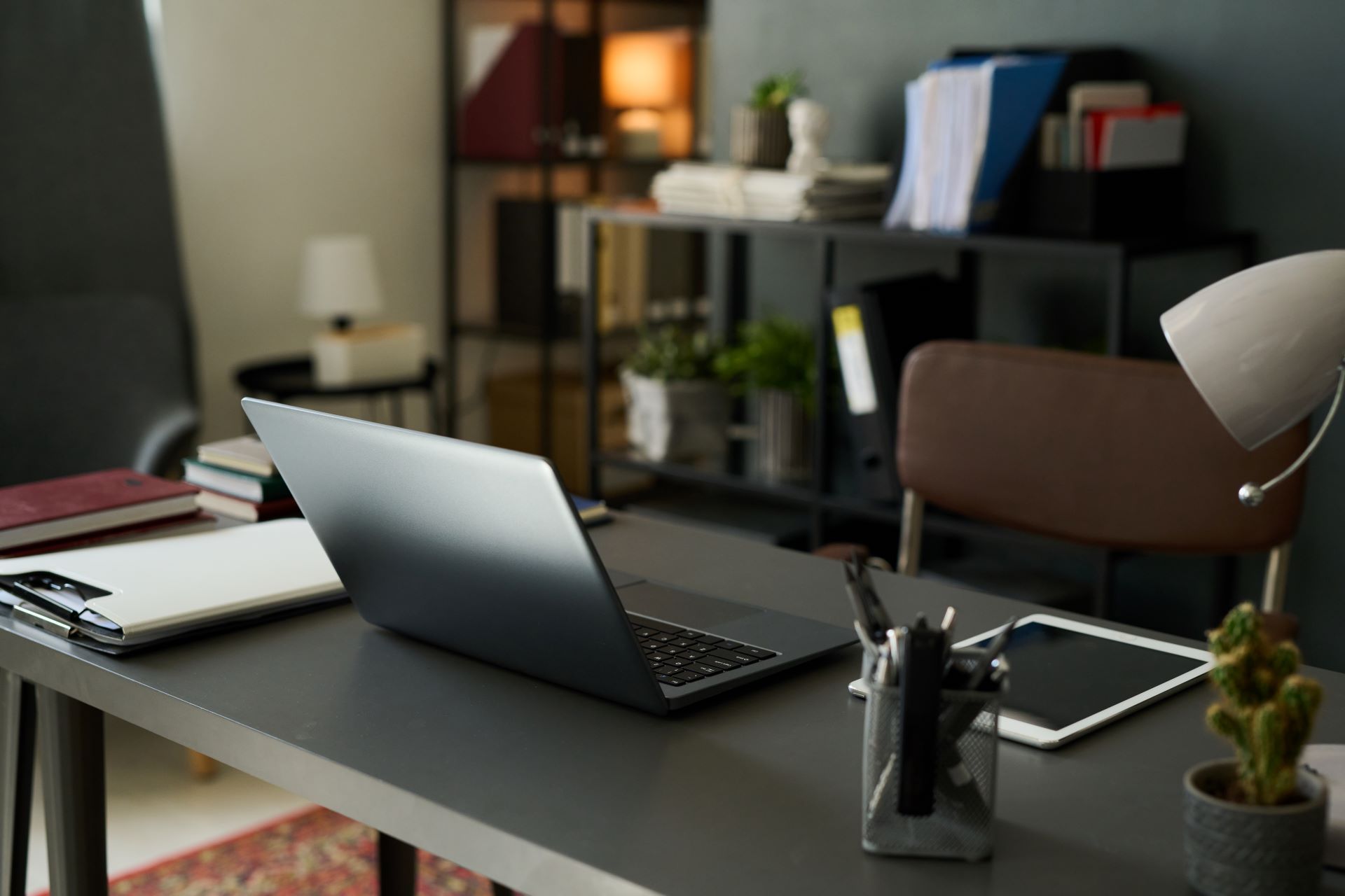 Laptop on a desk in a home office with a lamp, tablet, and plant.