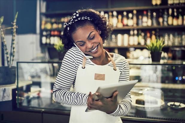 A smiling person in a striped shirt and apron stands in a cafe, talking on a phone while using a tablet.