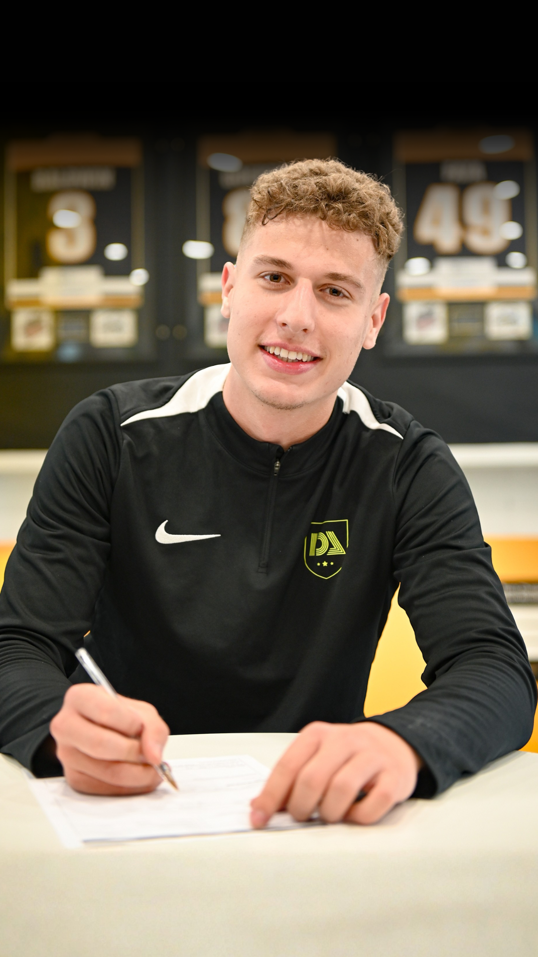 Man signing a document, wearing black athletic wear with logo. Background includes framed jerseys.