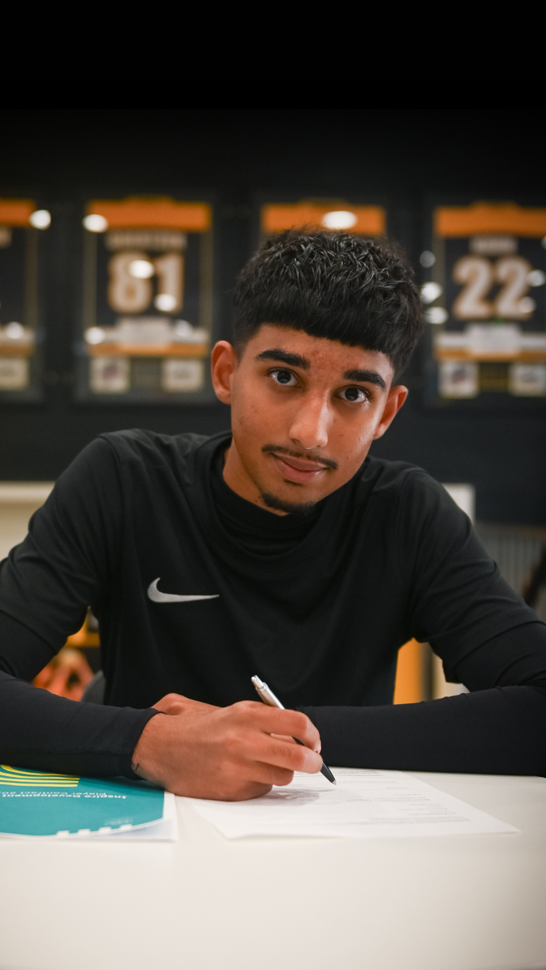 Person signing a document, wearing a black shirt, seated, with a neutral expression. Behind are framed jerseys.