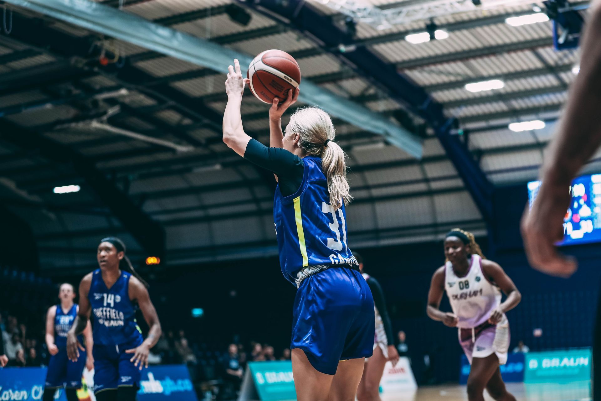 A basketball player in blue jersey shoots ball during game. Other players in the background.