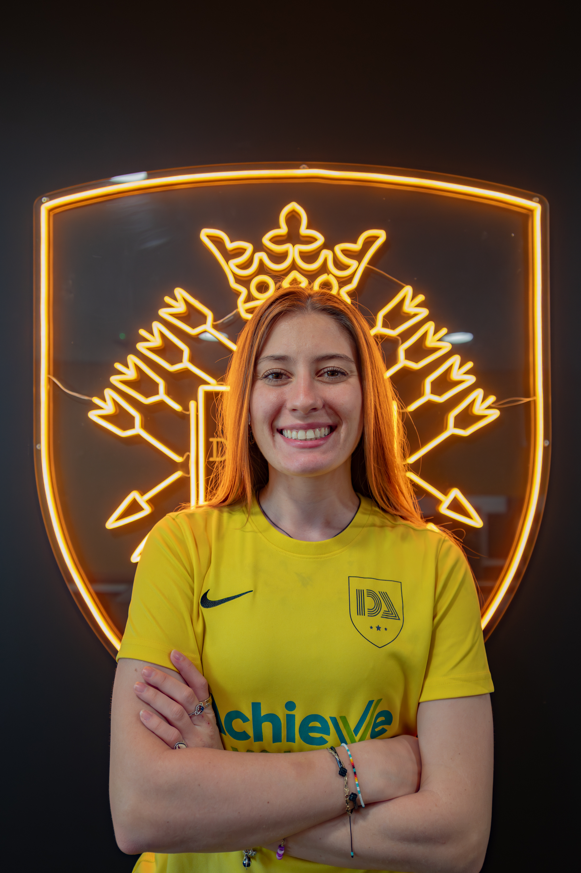 Woman in yellow jersey smiles, arms crossed, in front of a neon emblem.