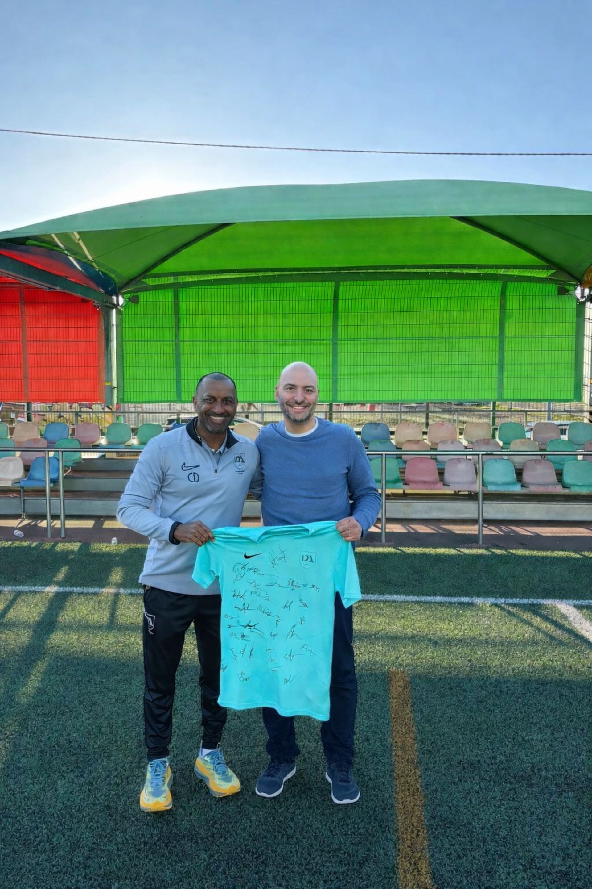 Two men on a soccer field holding a teal jersey, one smiles. Green and red bleachers in the background.