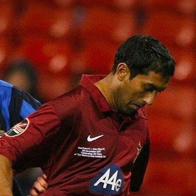 Soccer player in maroon jersey, head down, near another player with a blurred red background.